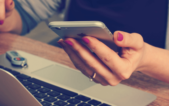 Close-up of a person's hands with pink nail polish holding a smartphone above a laptop keyboard, with a key fob beside the laptop.