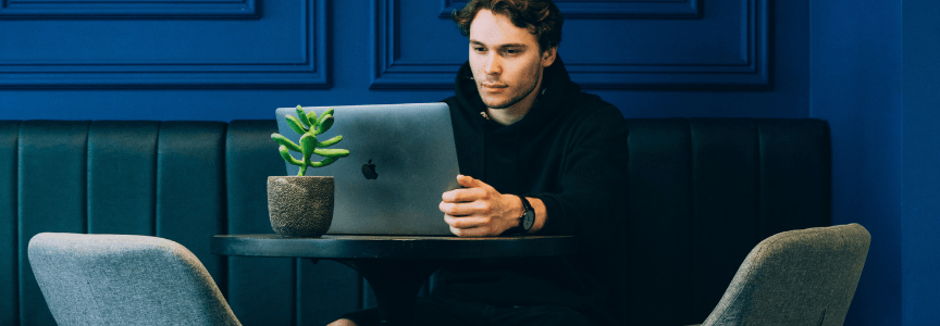 A man in a black turtleneck working on a laptop at a modern table with a small potted plant, seated on a dark green bench against a blue wall.