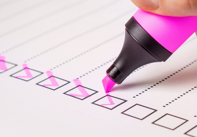 A hand holding a pink highlighter marking checkboxes on a printed checklist, with several boxes already checked.