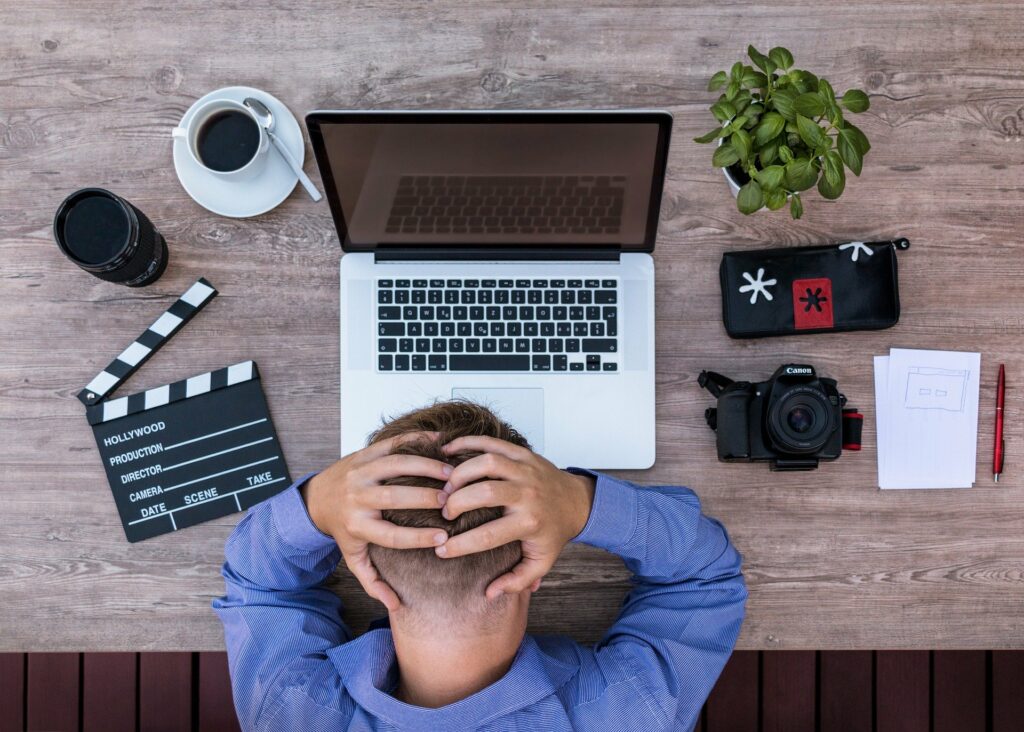 Overhead view of a person at a desk with their hands on their head in front of an open laptop, surrounded by creative work items including a coffee cup, camera, clapperboard, potted plant, and notes.