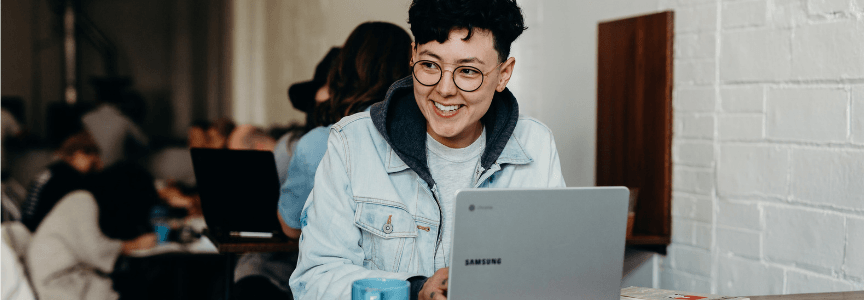 A person with glasses and a denim jacket smiles while working on a Samsung laptop in a busy indoor setting.