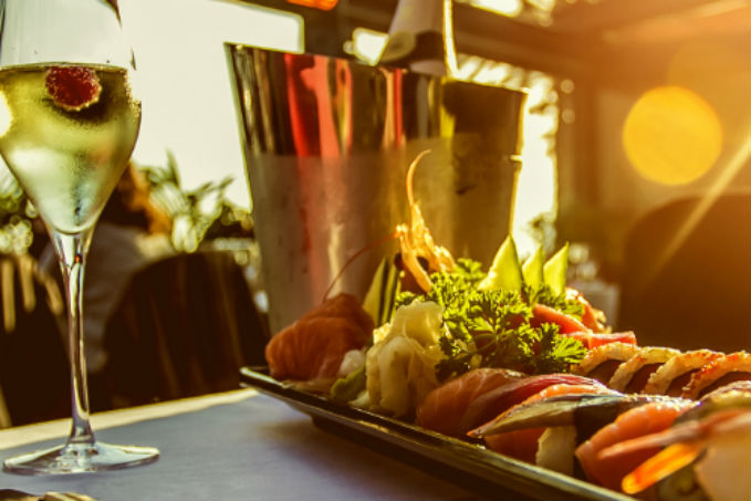 A plate of assorted sushi and sashimi with garnishes, a glass of white wine with a raspberry, and a wine cooler in an outdoor dining setting with warm sunlight.