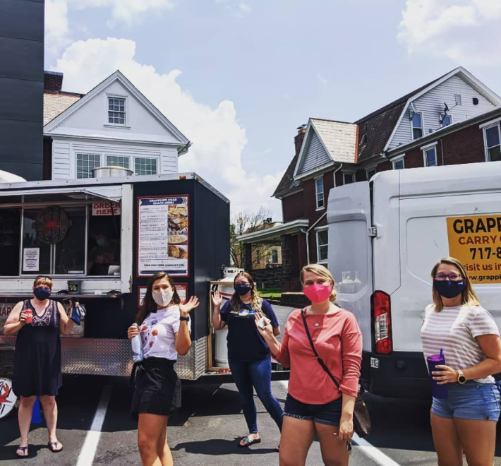 Four women wearing face masks standing in a parking lot in front of a food truck on a sunny day, with houses in the background.