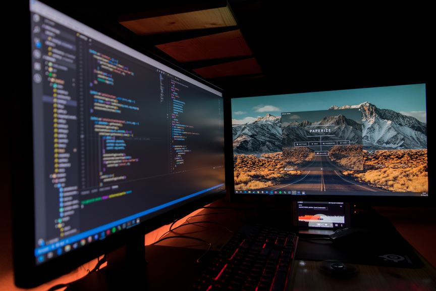 A dual monitor computer setup with a code editor on the left screen and a mountainous landscape wallpaper on the right. A keyboard and mouse are on the desk, indicating a programming workspace.