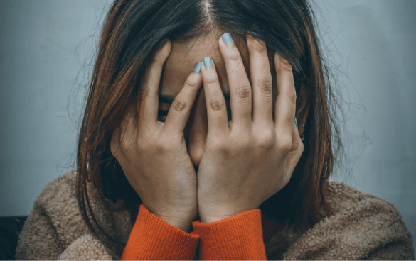 Person with brown hair wearing a red-orange sweater and beige jacket, covering their face with hands painted with light blue nail polish, against a gray background.