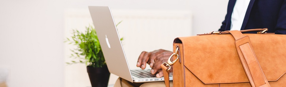 A person's hands typing on a MacBook with a tan leather briefcase and a potted plant on the desk, indicating a professional work environment.