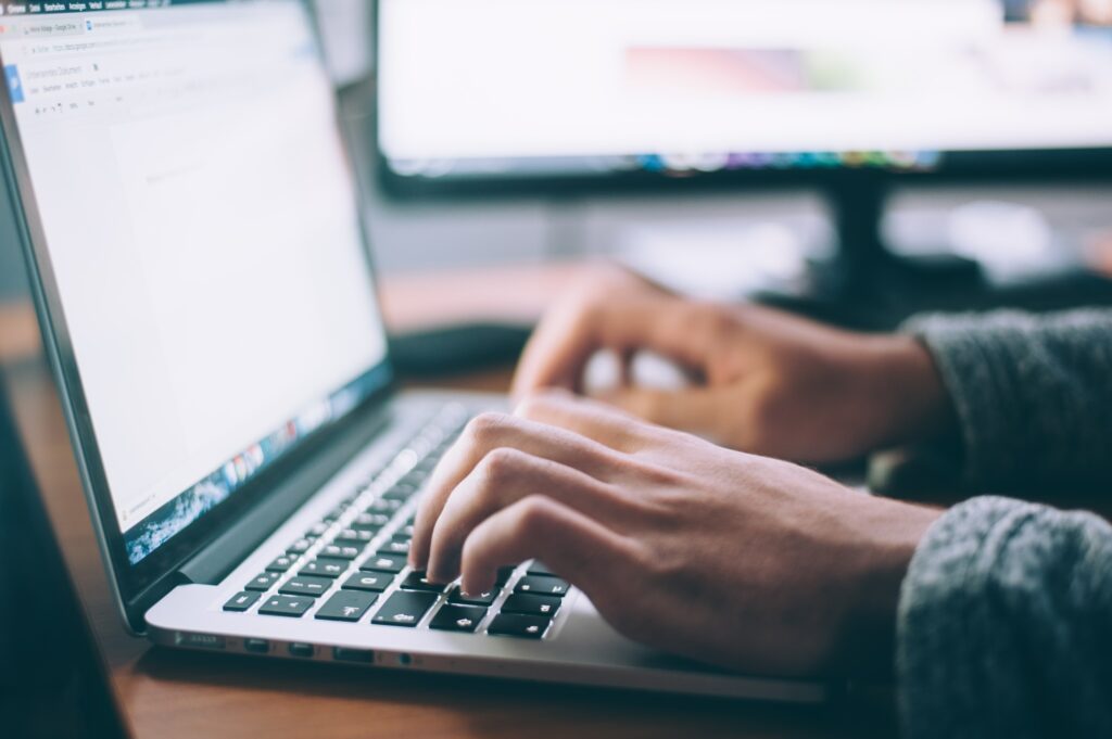 Close-up of a person's hands typing on a laptop keyboard with a blurred screen in the background, indicating a multi-screen workstation.