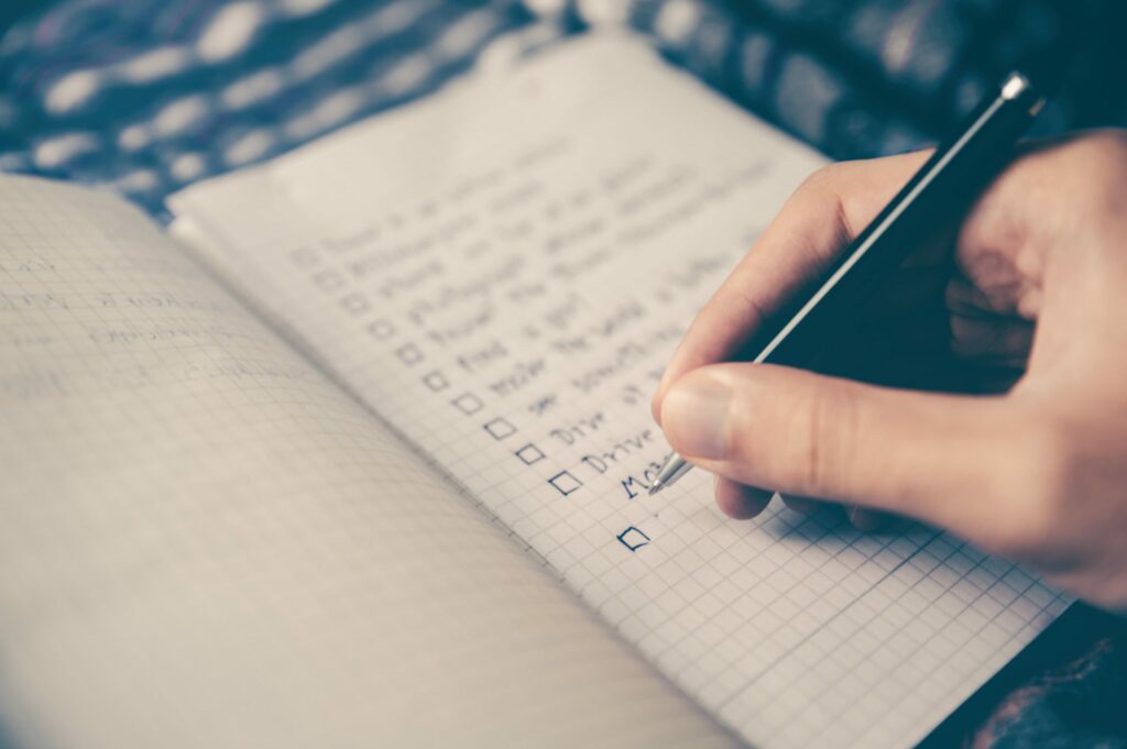 Close-up of a person's hand holding a pen and marking an item on a checklist in a notebook, with a patterned fabric in the blurred background.