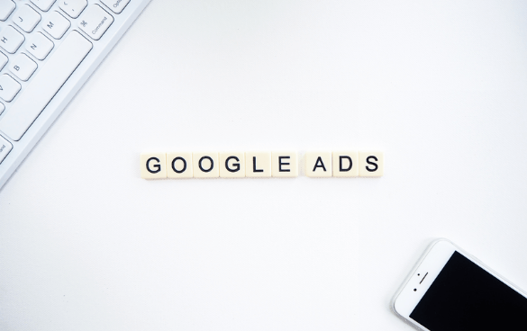 Scrabble-like tiles spelling 'GOOGLE ADS' on a white desk with a keyboard on the left and a smartphone on the right.