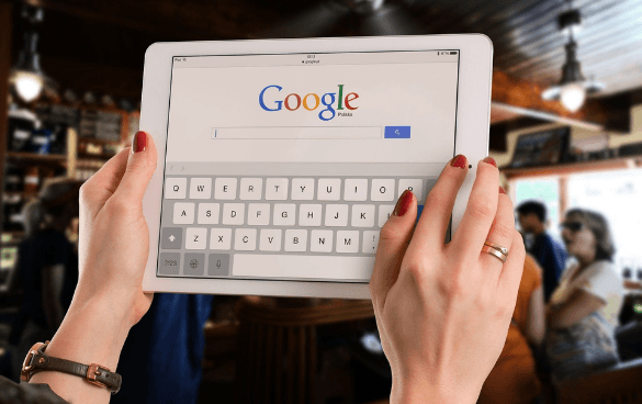 Person holding a tablet displaying Google search page with an on-screen keyboard, with a blurred cafe background.