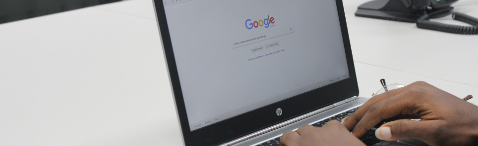 Person using an HP laptop with Google's homepage on the screen, with a white sheet of paper on the desk and a telephone in the background, indicating an office environment.