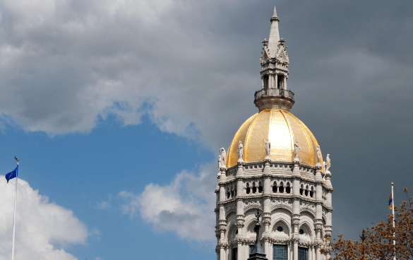 Close-up of a building's gold dome with ornate details, topped with a statue against a partly cloudy sky, and a blue flag on a flagpole to the left.