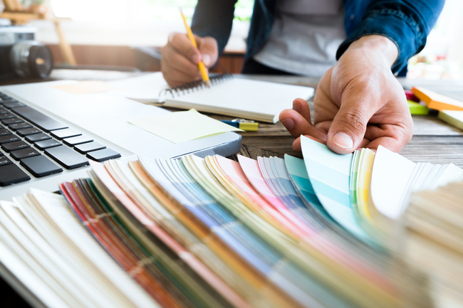 A person selecting color swatches with one hand and writing in a notebook with the other at a desk with a laptop, indicating a creative design process.