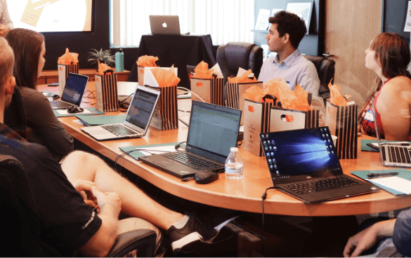 A group of people sitting around a conference table with laptops, listening to a presentation being given by a person at the head of the table.