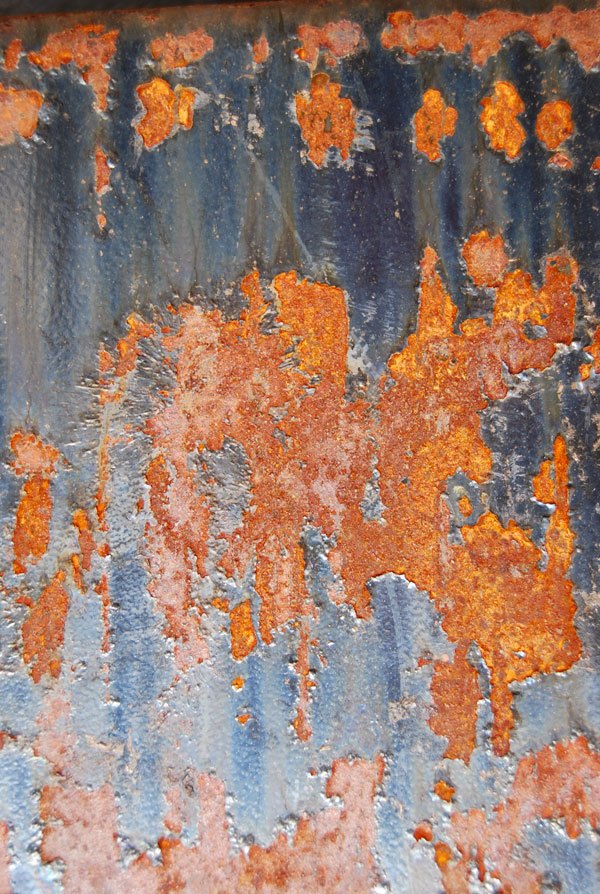 Close-up of a rusted metal surface with patches of orange and brown rust on the remaining blue-grey paint, showing a rough and flaky texture.