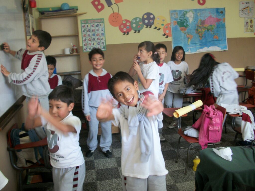 Energetic children in school uniforms playing and interacting in a lively classroom with educational posters and a world map on the wall.