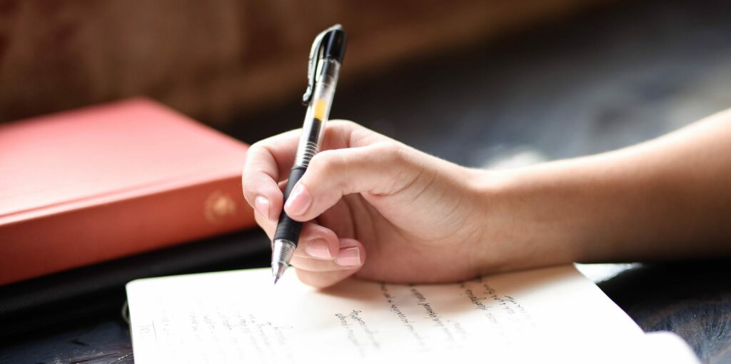 A person's hand writing in a notebook with a black and yellow pen, with a red book with a golden emblem in the blurred background.