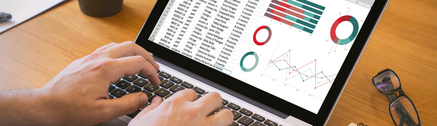 Person working on a laptop with spreadsheets and various charts displayed on the screen, indicating data analysis or financial work, with a pair of glasses and a coffee mug on the desk.