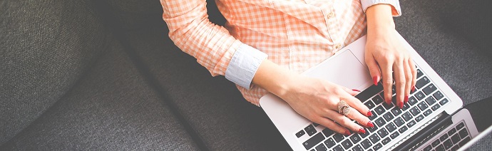 Person in a checkered shirt working on a laptop with hands on the keyboard, seated on a gray couch.