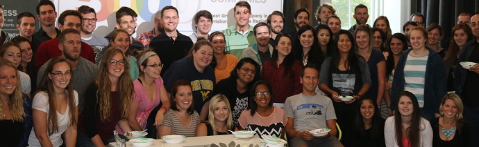 A diverse group of smiling people posing for a photo at a social or professional gathering, with some holding plates, suggesting the presence of food.