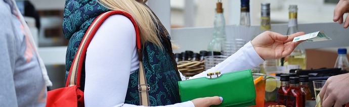 A person in a white top and patterned scarf is handing over cash from a green wallet to another person, with bottles in the background indicating a market or store setting.