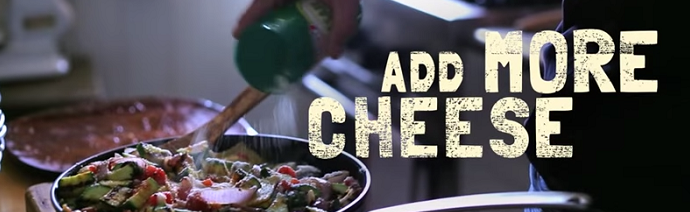 A person is pouring grated cheese from a green bottle onto a dish of salad or mixed vegetables, with the phrase 'AND MORE CHEESE' displayed at the top.