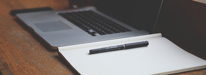 A partially closed laptop on a wooden surface with an open blank notebook and a black pen in front.