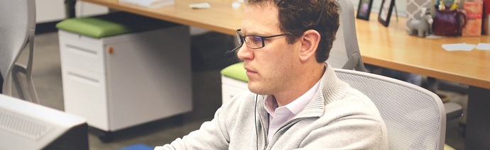 A man wearing glasses and a grey cardigan working intently at a computer in an office setting.