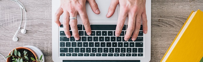 Person typing on a laptop with a small potted plant, white earbuds, and a closed yellow notebook on a wooden surface.