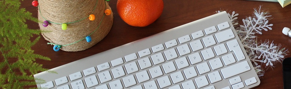 A festive workspace with a computer keyboard, a spool wrapped with string lights, a tangerine, a snowflake decoration, and a small evergreen branch.