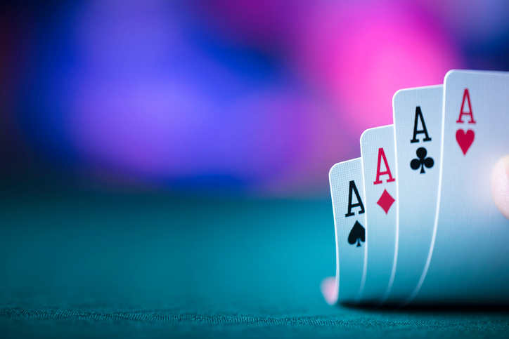 Four aces from a standard deck of playing cards are fanned out on a table with a blurred purple and blue bokeh background.