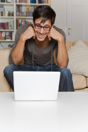 A cheerful man with glasses sitting at a table with a laptop, fists clenched near his cheeks in excitement, with a couch and bookshelf in the background.