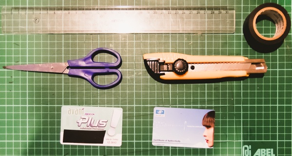 A pair of scissors, two plastic cards, a rotary cutter, and a roll of tape on a green cutting mat with grid lines.