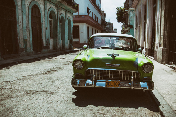 A vintage green car parked on a cobblestone street with old buildings on either side, under a bright sky.