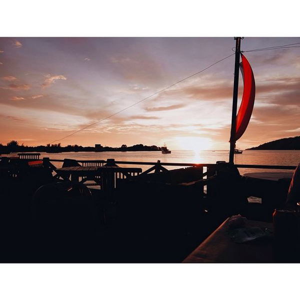 Sunset over water with a sailboat featuring a red and black sail in the foreground, and outdoor furniture silhouetted against the warm sky.