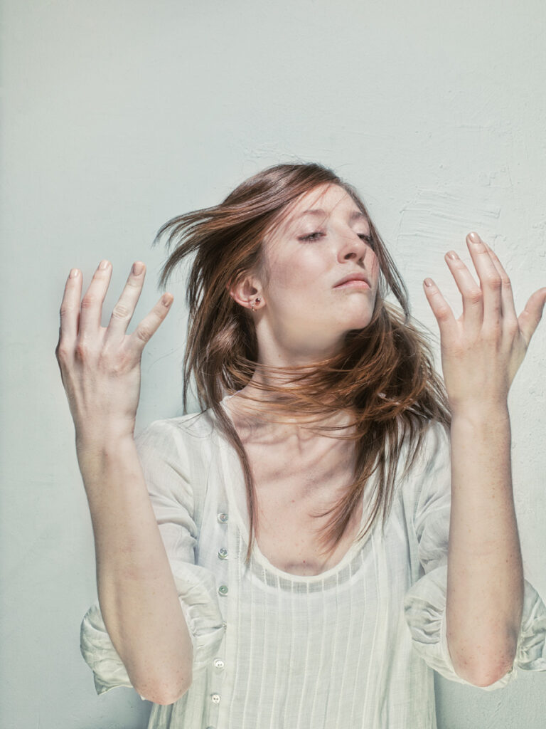 A woman with closed eyes, head tilted back, and windswept hair, wearing a light blouse with raised arms and open hands, conveying a sense of feeling the air or expressing freedom.