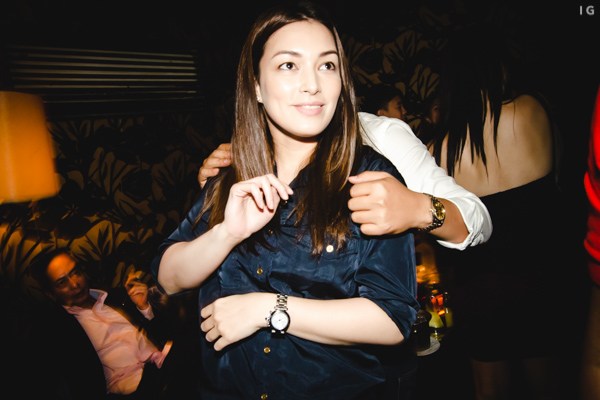 A smiling woman with long hair wearing a dark blue shirt and a watch, dancing or moving in a dimly lit social setting with patterned wallpaper and blurred figures of other people in the background.