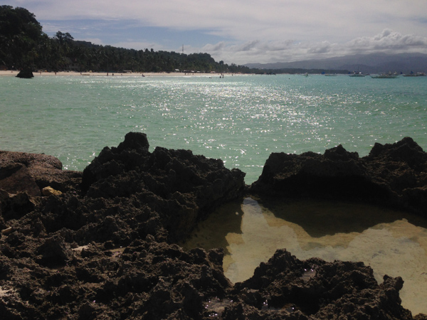 Rugged rocks in the foreground with a small pool of water, overlooking a tropical beach with sparkling turquoise waters, white sand, lush greenery, and boats in the distance under a sunny sky.