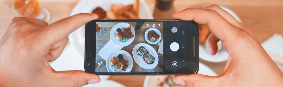 Hands holding a smartphone taking a photo of various dishes on a table.