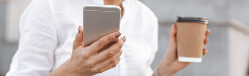 Close-up of a person's hands holding a smartphone and a takeaway coffee cup, wearing a white shirt, with a blurred background.