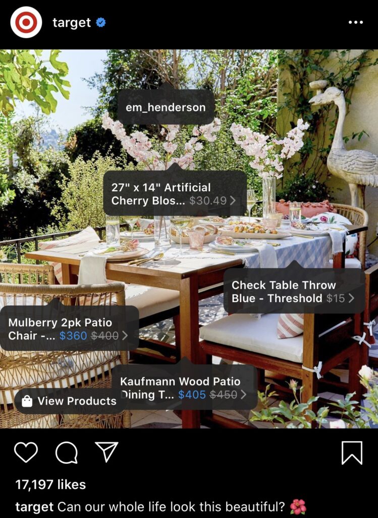 Outdoor dining area set for a meal with a blue checkered tablecloth, wooden chairs, artificial cherry blossoms, and a bird sculpture, surrounded by greenery.