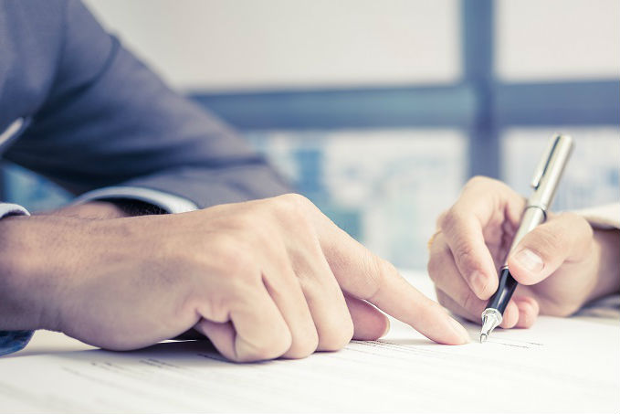 Close-up of a person's hands with one pointing to a line on a document and the other holding a pen, ready to write, with a blurred office background.