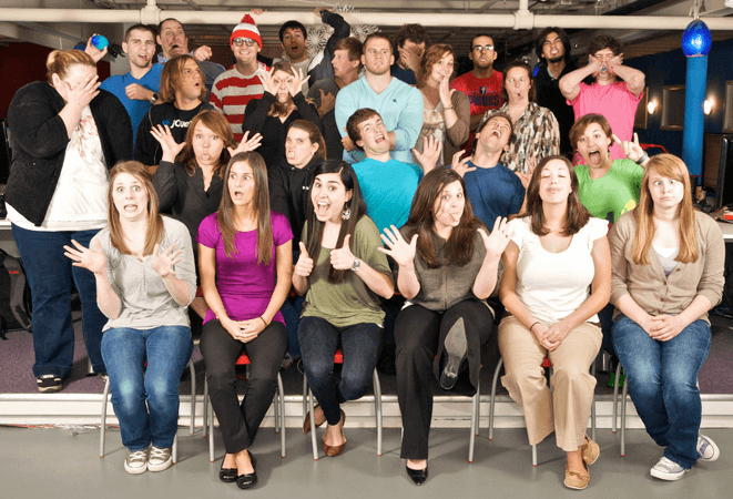 A diverse group of people posing with a range of expressions from surprise to laughter, with one individual dressed as Waldo from 'Where's Waldo?' in the background.