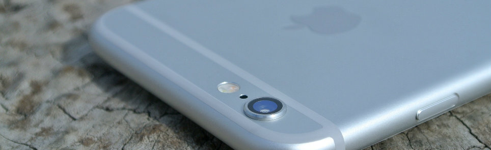 Close-up of the upper back of a silver iPhone showing the camera lens, on a textured surface resembling cracked earth.