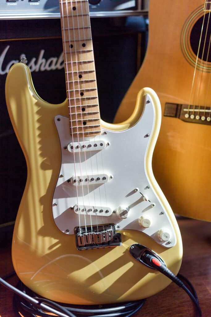 A yellow electric guitar with a white pickguard in front of a Marshall amplifier, with a cable plugged in and part of an acoustic guitar visible to the right.