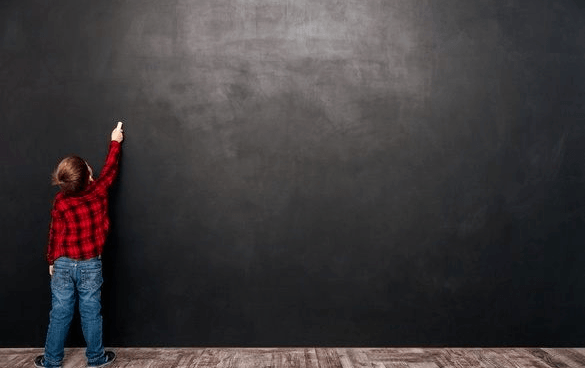 A young child in a red and black plaid shirt and blue jeans is reaching up to a large, dark chalkboard, appearing to write or draw.