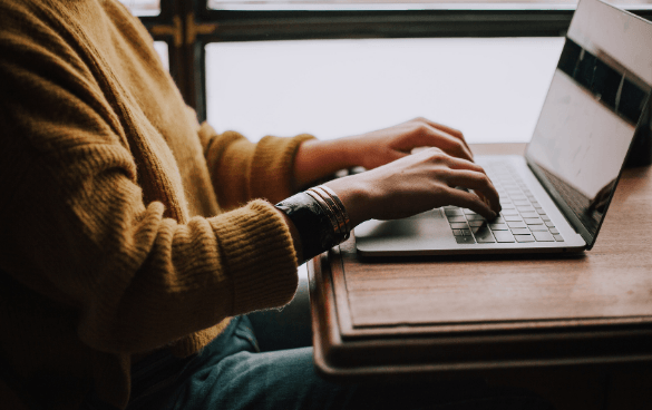 Person in a yellow sweater typing on a laptop at a wooden table, with a bracelet on their wrist and a blurred background suggesting a bright interior space.