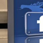 Three-dimensional Facebook logo in front of a world map, with a partial view of a Facebook interface on a computer screen to the left.