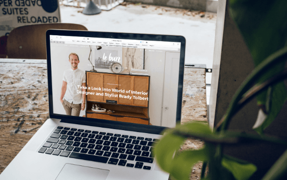 An open laptop on a table displaying a website featuring an article about interior designer and stylist Brady Tolbert, with his photo next to a wooden cabinet.