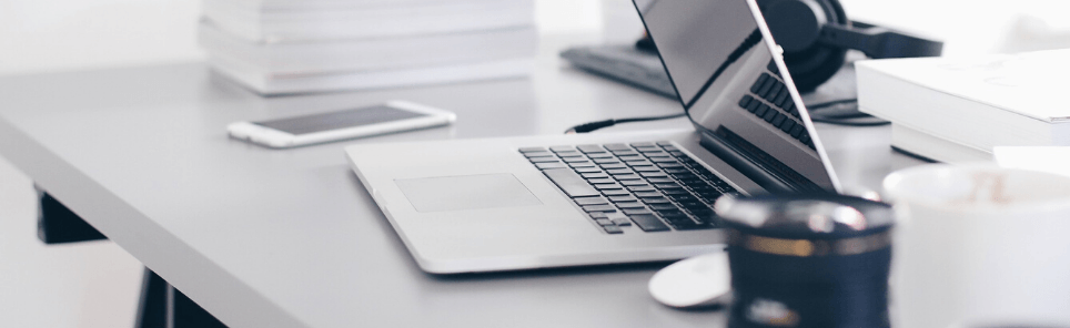 A tidy workspace with a laptop, smartphone, stack of books, closed notebook, headphones, and a cup of coffee on a white desk.